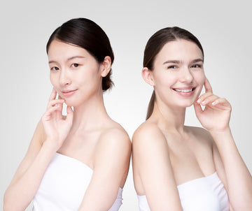 Two women stand back-to-back, smiling gently. Both touch their faces, conveying elegance and freshness.