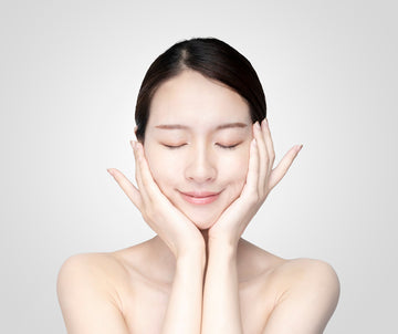 A woman with closed eyes and a serene smile gently touches her cheeks with both hands. Her hair is neatly tied back, and her skin appears smooth and glowing.